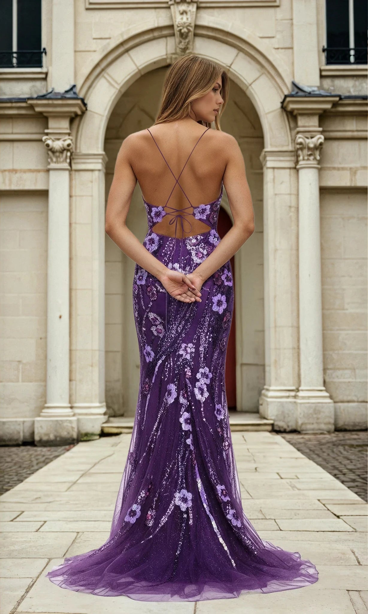 A woman poses with her back to the camera in the Floral Sequin-Print Long Purple Prom Dress L569, which features crisscross straps and an open back, in front of a light stone building with arched doorways.